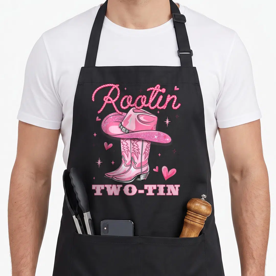 Rootin cowgirl aprons - Pink cowboy hat on boots with rope text reading Rootin Two-Tin and scattered hearts — shown on a black cotton canvas apron against a white background
