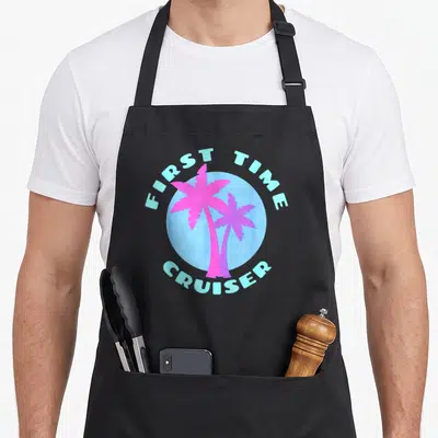Cruise cruiser girl apron - Two pink palm trees in light blue circle with FIRST TIME CRUISER text — shown on a black cotton canvas apron against a white background