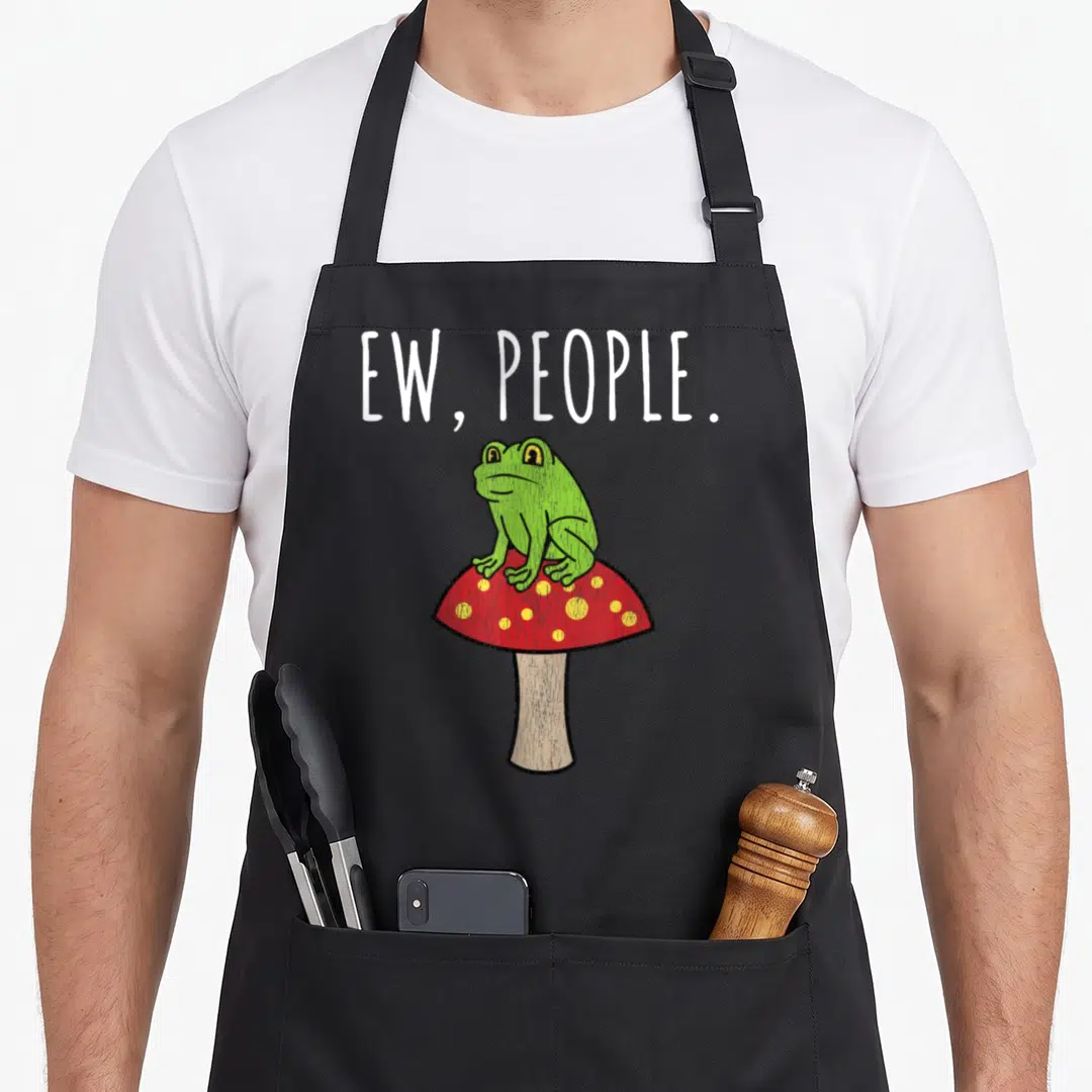 Funny frog mushroom apron - Green frog sitting on red mushroom with yellow dots under white EW, PEOPLE text — shown on a black cotton canvas apron against a white background