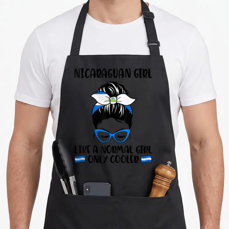 Normal nicaraguan girl apron - Cartoon girl with blue glasses and Nicaragua flag headband, text reads NICARAGUAN GIRL — shown on a black cotton canvas apron against a white background