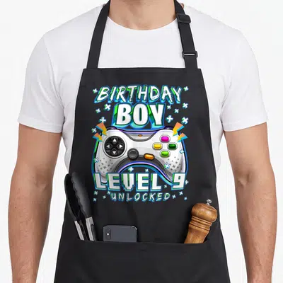 Gamer age video game apron - Cartoon controller with BIRTHDAY BOY LEVEL 9 UNLOCKED text and geometric shapes — shown on a black cotton canvas apron against a white background