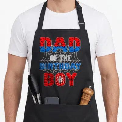 Spider birthday apron - Stacked DAD OF THE BIRTHDAY BOY text in red and blue with spider web pattern and white spider — shown on a black cotton canvas apron against a white background