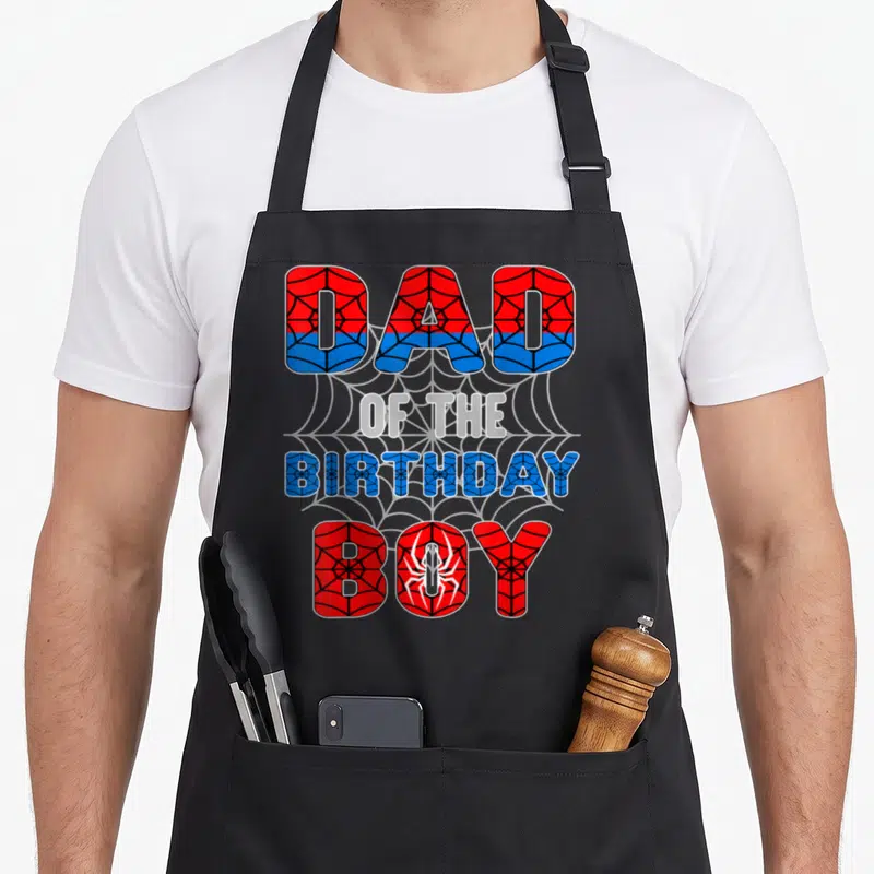 Spider birthday apron - Stacked DAD OF THE BIRTHDAY BOY text in red and blue with spider web pattern and white spider — shown on a black cotton canvas apron against a white background