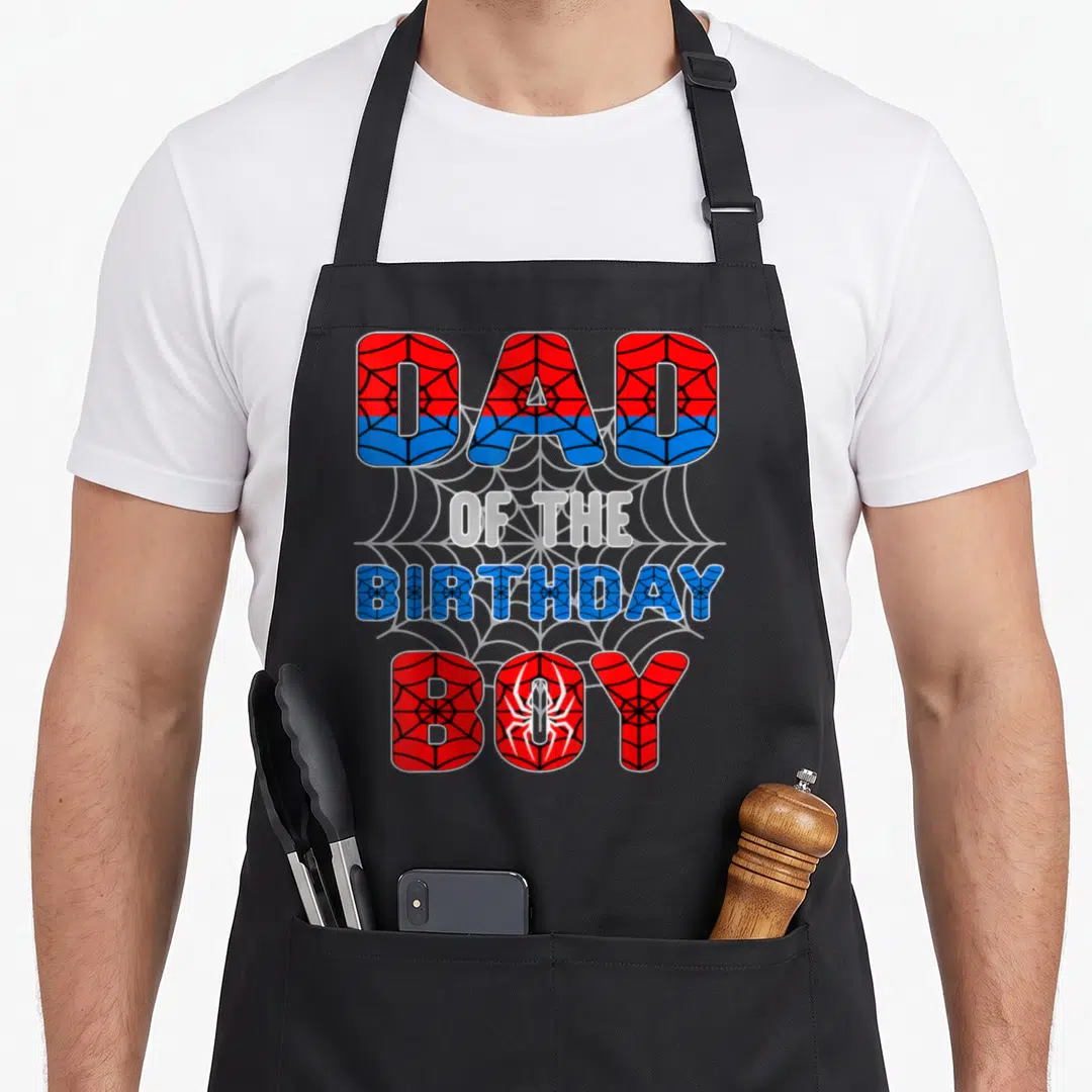 Spider birthday apron - Stacked DAD OF THE BIRTHDAY BOY text in red and blue with spider web pattern and white spider — shown on a black cotton canvas apron against a white background
