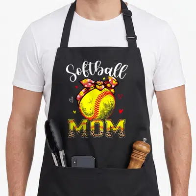 Headband leopard print cooking apron - Softball MOM text with yellow ball, red bow, hearts, and leopard print letters — shown on a black cotton canvas apron against a white background