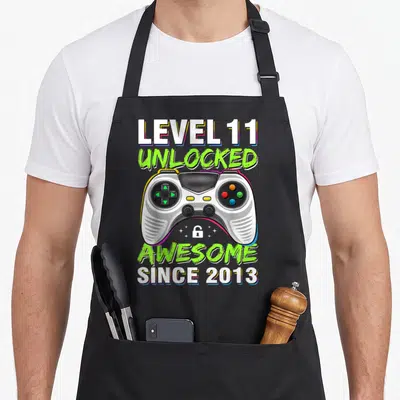 Level unlocked 11th gaming aprons - Game controller with LEVEL 11 UNLOCKED AWESOME SINCE 2013 text and lock icon — shown on a black cotton canvas apron against a white background