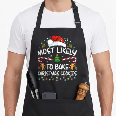 Likely women's christmas aprons - Santa hat and gingerbread men surrounding MOST LIKELY TO BAKE CHRISTMAS COOKIES text — shown on a black cotton canvas apron against a white background