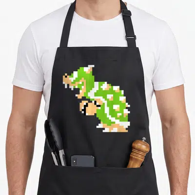 Bit holiday apron - Green pixel art Koopa Troopa sprite centered on canvas — shown on a black cotton canvas apron against a white background