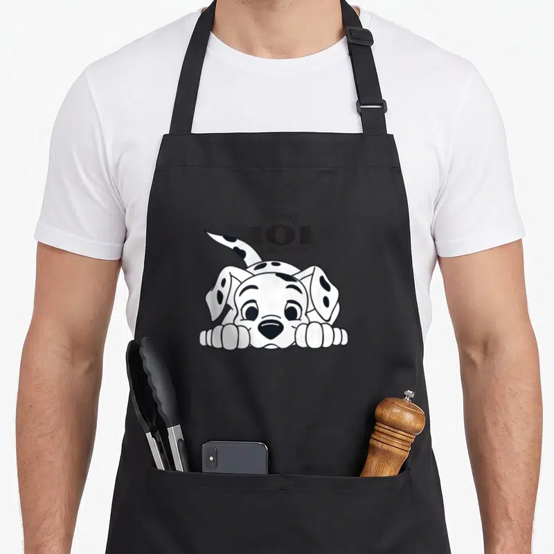 Playful dalmatian apron - Disney 101 Dalmatians text above a cartoon puppy with black spots resting on paws — shown on a black cotton canvas apron against a white background