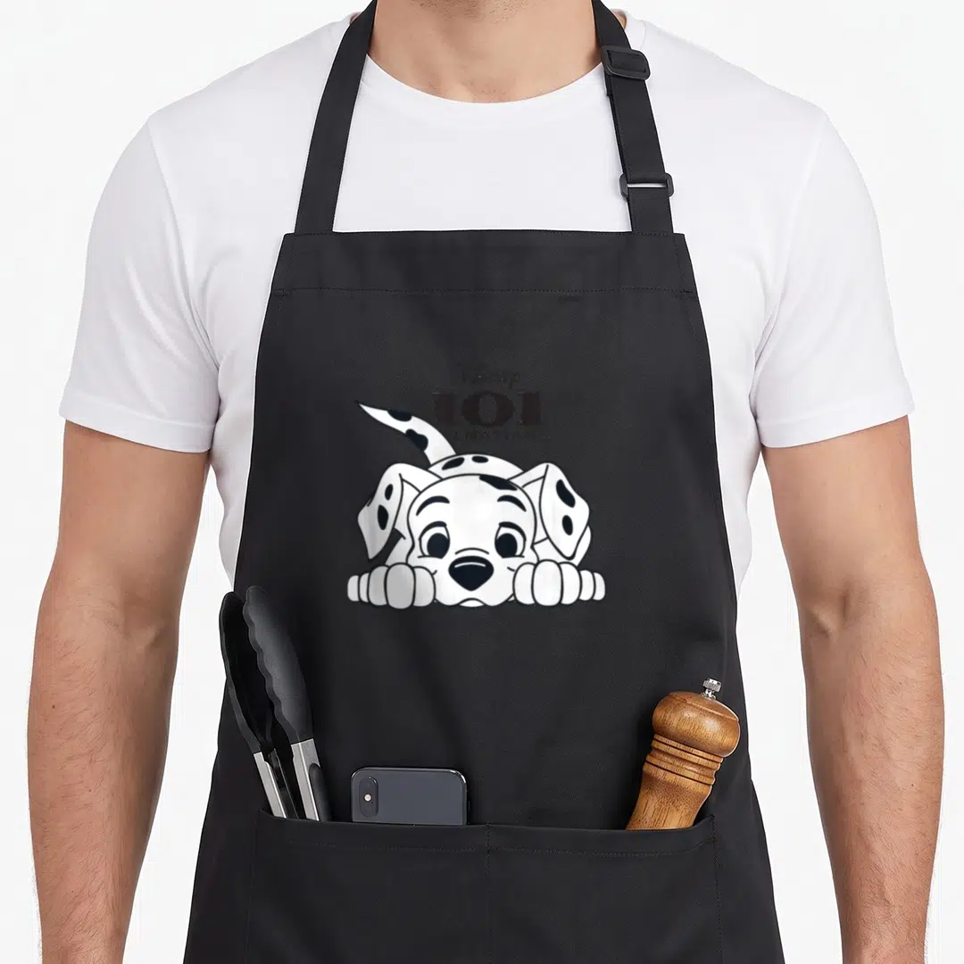 Playful dalmatian apron - Disney 101 Dalmatians text above a cartoon puppy with black spots resting on paws — shown on a black cotton canvas apron against a white background
