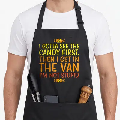 Van halloween apron - Stacked yellow orange red text reading I Gotta See Candy First Van flanked by candy icons — shown on a black cotton canvas apron against a white background