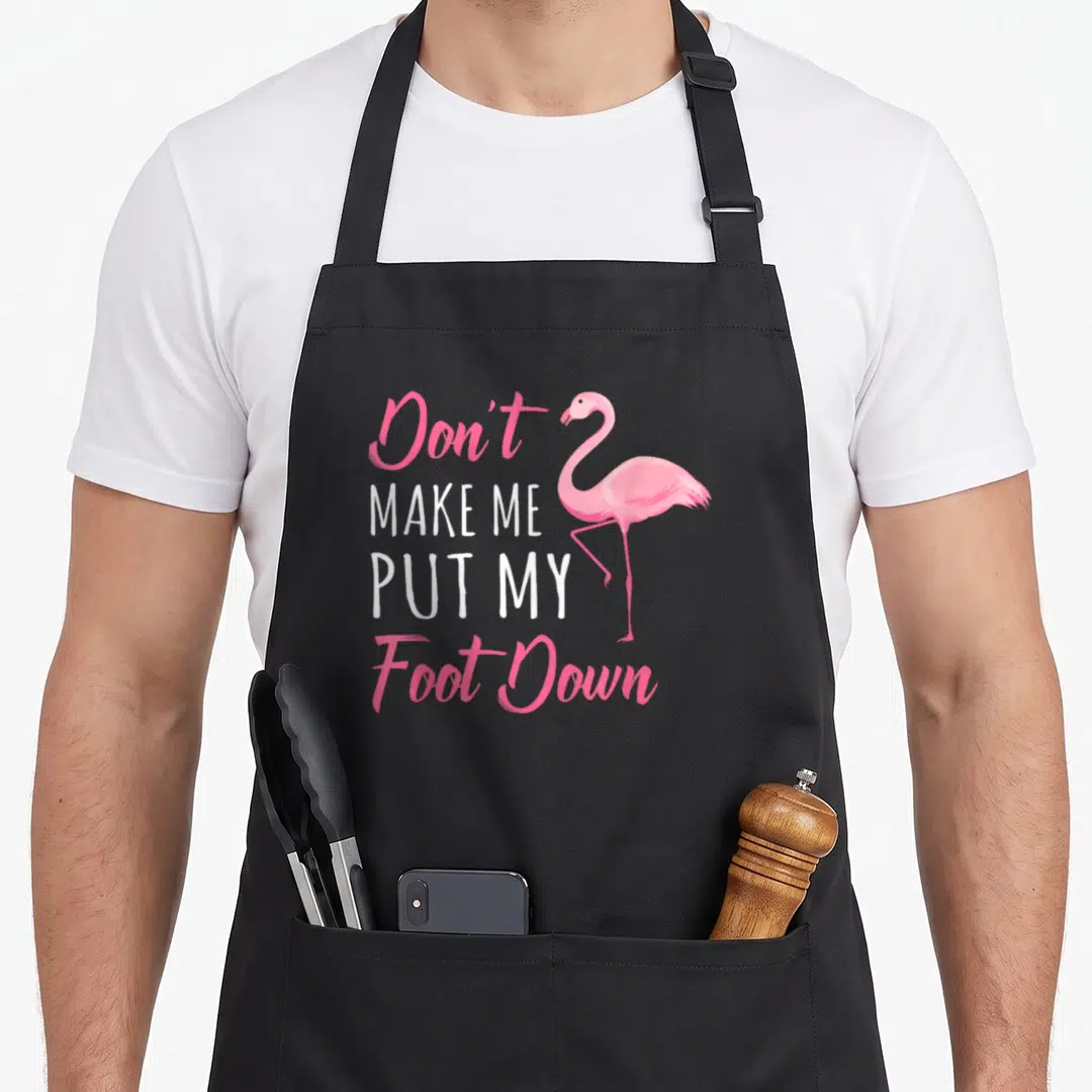 Cute foot flamingo apron - Pink flamingo stands on one leg beside stacked text reading Don't Make Me Put My Foot Down — shown on a black cotton canvas apron against a white background