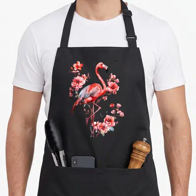 Motif the floral apron - Watercolor pink flamingo standing among red and blue floral branches with small butterflies — shown on a black cotton canvas apron against a white background