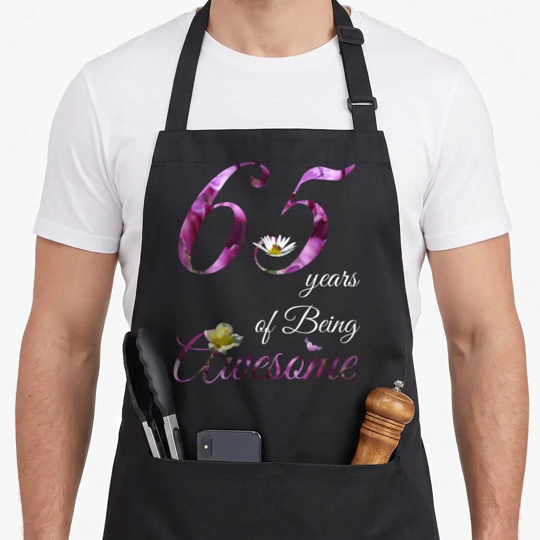 65th floral aprons - Purple floral 65 with daisy bee and yellow rose typography — shown on a black cotton canvas apron against a white background