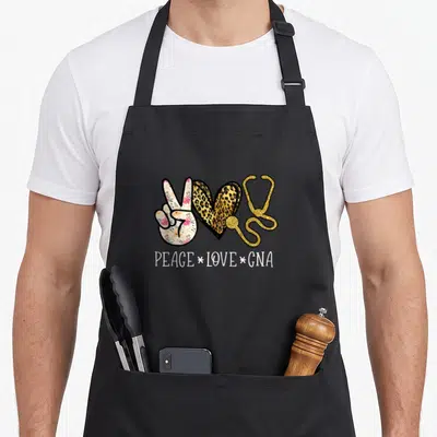 Nurselife nurse apron - Floral peace sign, leopard heart, gold stethoscope, and PEACE LOVE GNA text — shown on a black cotton canvas apron against a white background