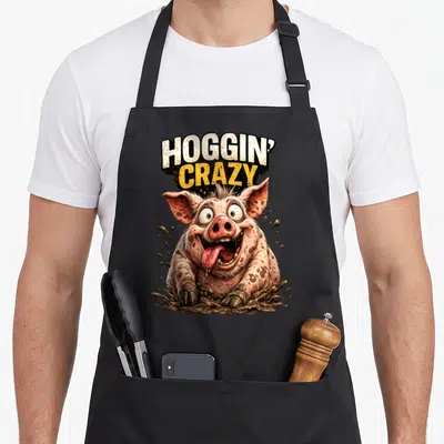 Hoggin crazy pig man apron - Cartoon pig emerging from brown mud under stacked HOGGIN' CRAZY text in white and yellow — shown on a black cotton canvas apron against a white background