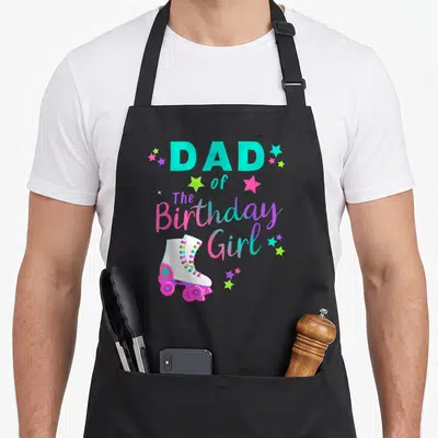 Skate family aprons - DAD of The Birthday Girl text with cartoon roller skate and multicolored stars — shown on a black cotton canvas apron against a white background