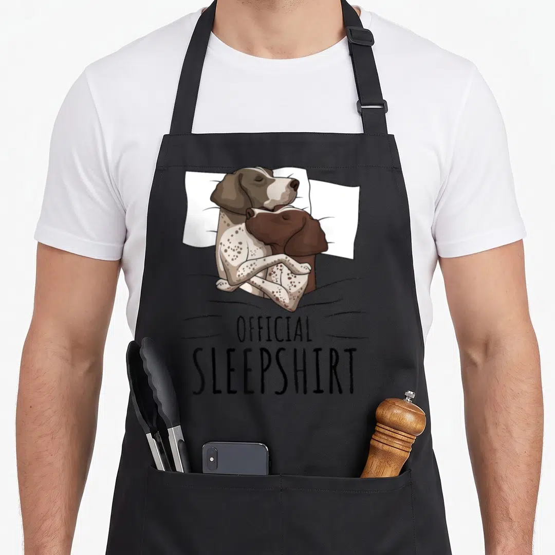 German shorthaired dog apron - Two cartoon dogs cuddling on a pillow under a grey blanket above official sleepshirt text — shown on a black cotton canvas apron against a white background