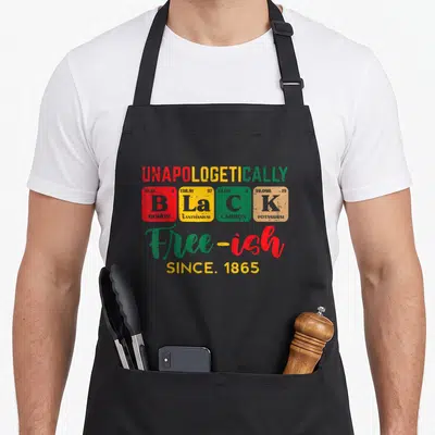 Unapologetically black apron - Stacked text with four colored element squares spelling BLACK and Free-ish since 1865 — shown on a black cotton canvas apron against a white background