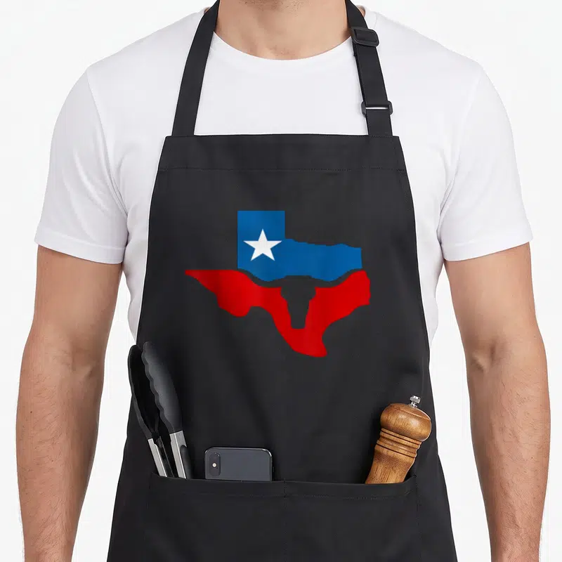 Longhorn texas apron - Texas state map silhouette with white star and bull horn cutout in blue red and white — shown on a black cotton canvas apron against a white background