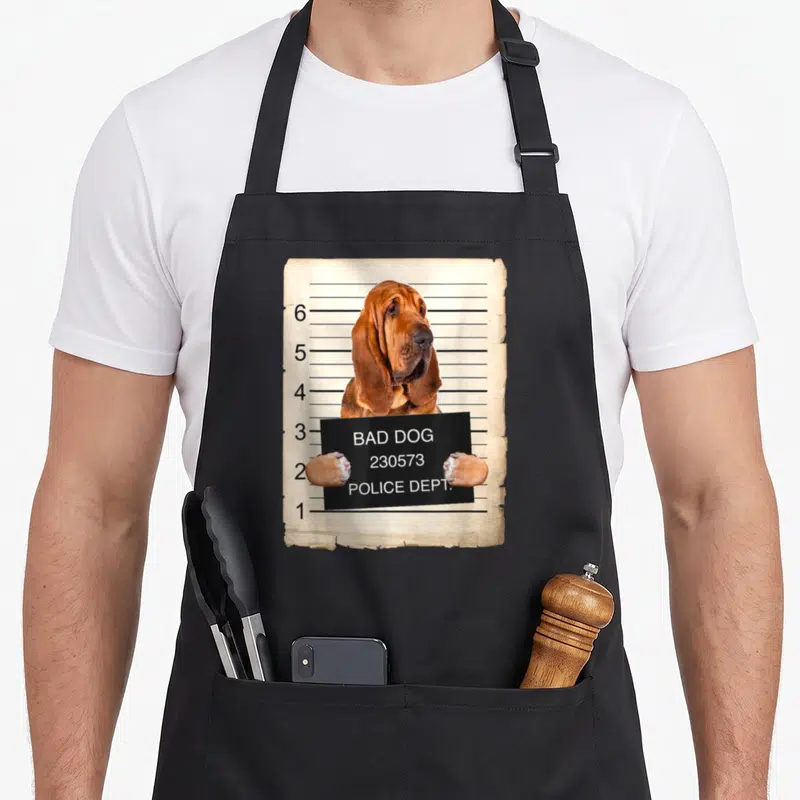 Bloodhound mug dog apron - Brown bloodhound holding a black BAD DOG POLICE DEPT sign against a beige height chart — shown on a black cotton canvas apron against a white background