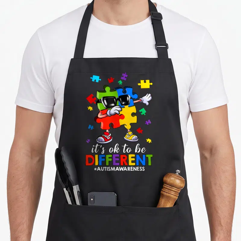 Different autism awareness apron - Cartoon puzzle piece character with sunglasses and text it's ok to be DIFFERENT — shown on a black cotton canvas apron against a white background