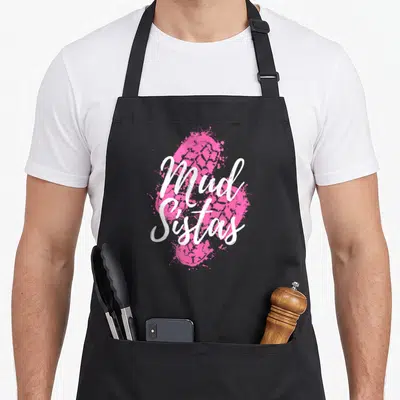 Sistas mud apron - White Mud Sistas text over pink tire track and paint splatter art — shown on a black cotton canvas apron against a white background