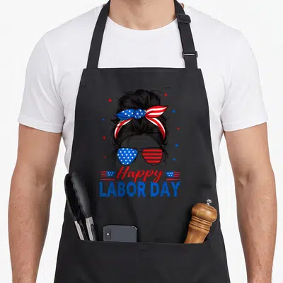 Labor messy aprons - Cartoon woman with messy bun, patriotic sunglasses, and Happy Labor Day text in red white blue — shown on a black cotton canvas apron against a white background