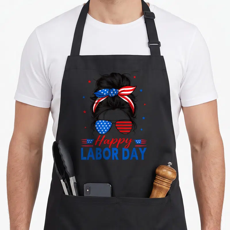 Labor messy aprons - Cartoon woman with messy bun, patriotic sunglasses, and Happy Labor Day text in red white blue — shown on a black cotton canvas apron against a white background