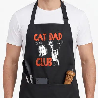 Cute better cat apron - Orange CAT DAD CLUB text with two white cartoon cats and script slogan — shown on a black cotton canvas apron against a white background