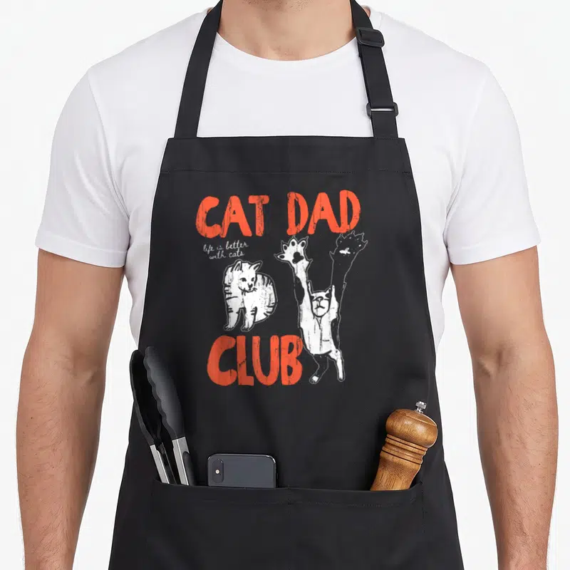 Cute better cat apron - Orange CAT DAD CLUB text with two white cartoon cats and script slogan — shown on a black cotton canvas apron against a white background