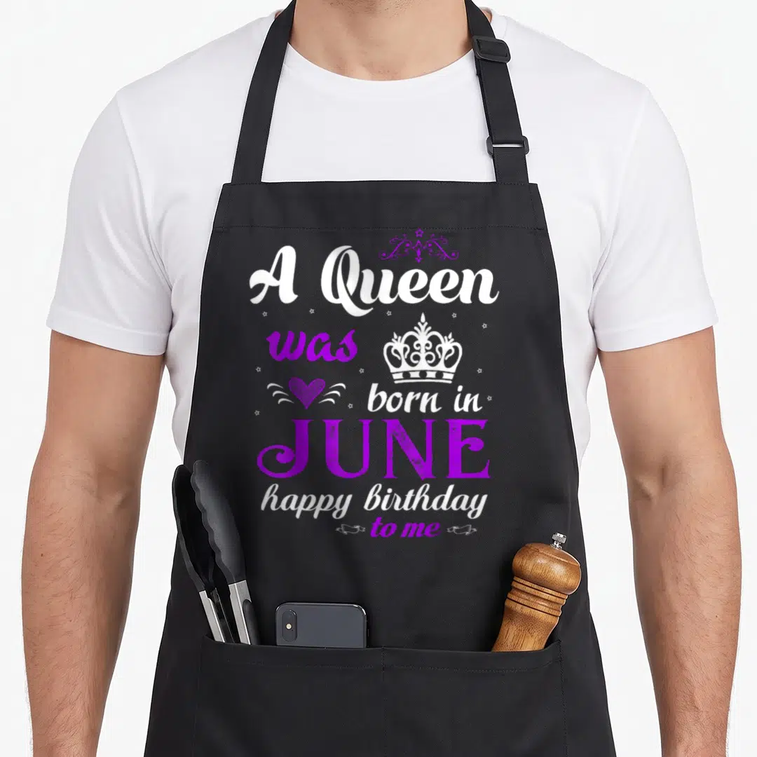 Queen june birthday apron - Stacked white and purple text A Queen was born in JUNE with crown heart and stars — shown on a black cotton canvas apron against a white background