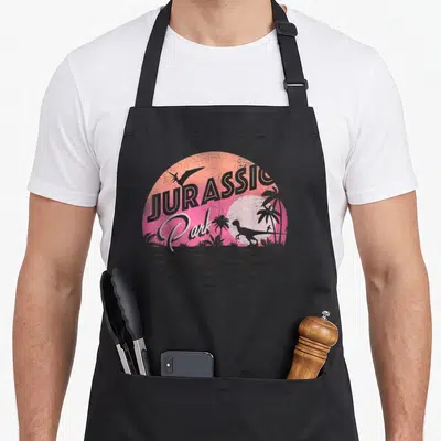90s dinosaur apron - JURASSIC Park text over pink orange sun with dinosaur palm tree silhouettes and horizontal lines — shown on a black cotton canvas apron against a white background