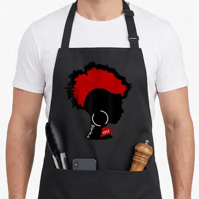 Dst tattoo apron - Black woman silhouette with red hair, vertical Divine text, and red 1913 triangle earring — shown on a black cotton canvas apron against a white background