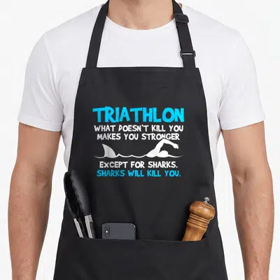 Funny triathlete triathlon apron - Blue and white stacked text with swimmer arm and shark fin graphic — shown on a black cotton canvas apron against a white background