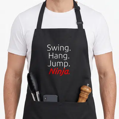 Swing jump apron - Stacked white and red text reading Swing Hang Jump Ninja on black fabric — shown on a black cotton canvas apron against a white background