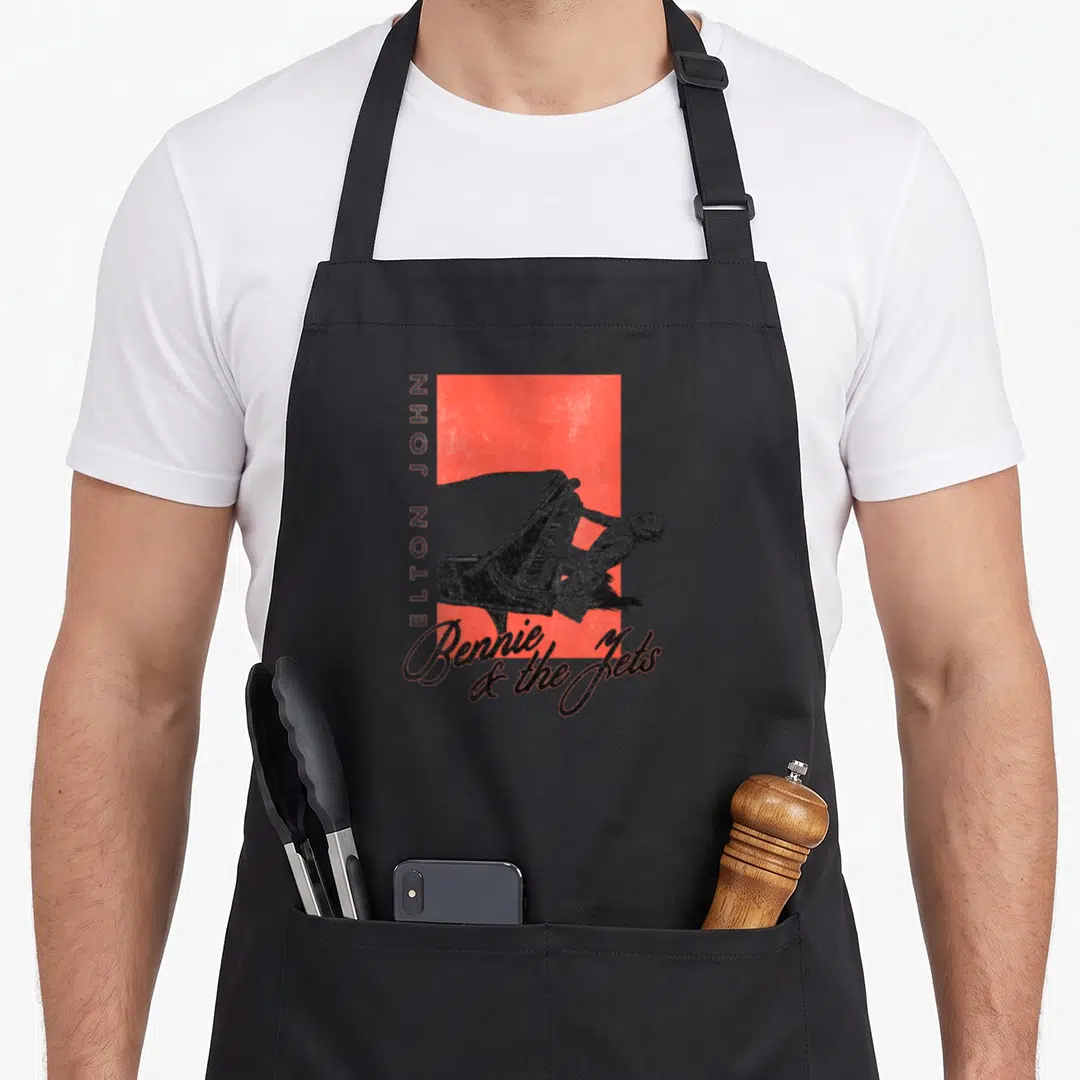 Bennie piano apron - Red rectangle with man leaning on piano, vertical ELTON JOHN text, and Bennie & the Jets script — shown on a black cotton canvas apron against a white background