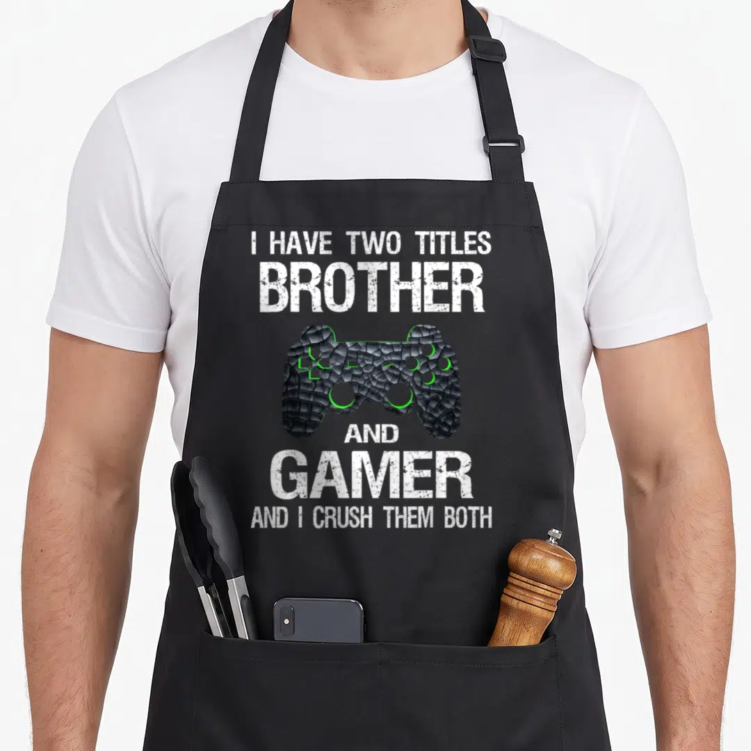 Gamer gaming video game apron - Distressed white text around black cracked game controller with green accents — shown on a black cotton canvas apron against a white background