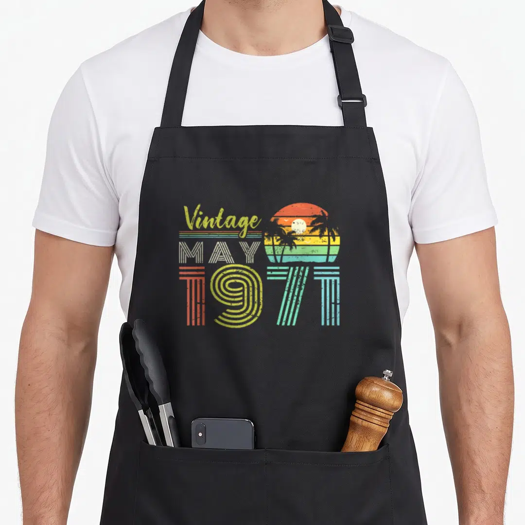 50th may birthday apron - Retro sunset with palm trees and birds beside striped Vintage MAY 1971 text in orange yellow — shown on a black cotton canvas apron against a white background