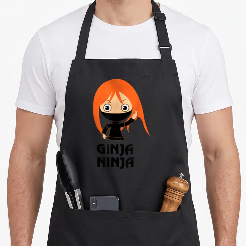 Ginja hair apron - Cartoon ninja with orange hair above GINJA NINJA text — shown on a black cotton canvas apron against a white background