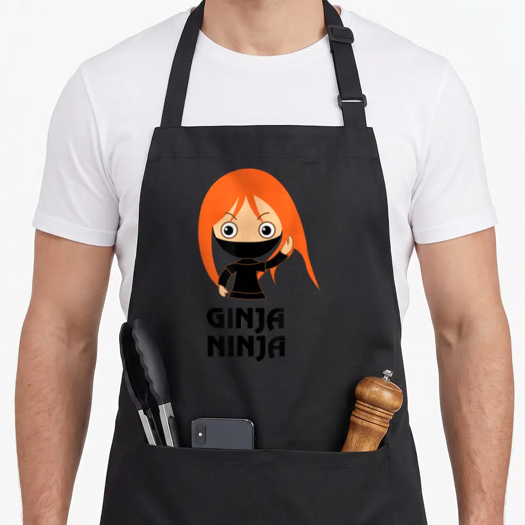 Ginja hair apron - Cartoon ninja with orange hair above GINJA NINJA text — shown on a black cotton canvas apron against a white background