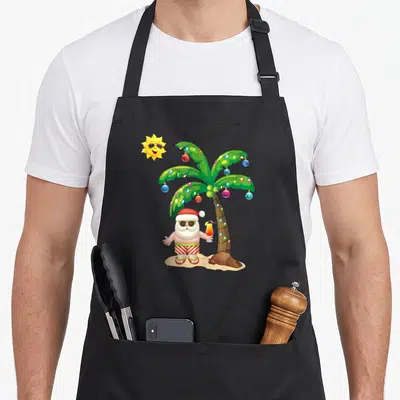 July santa claus apron - Cartoon Santa in red shorts holding a cocktail under a green palm tree with ornaments — shown on a black cotton canvas apron against a white background