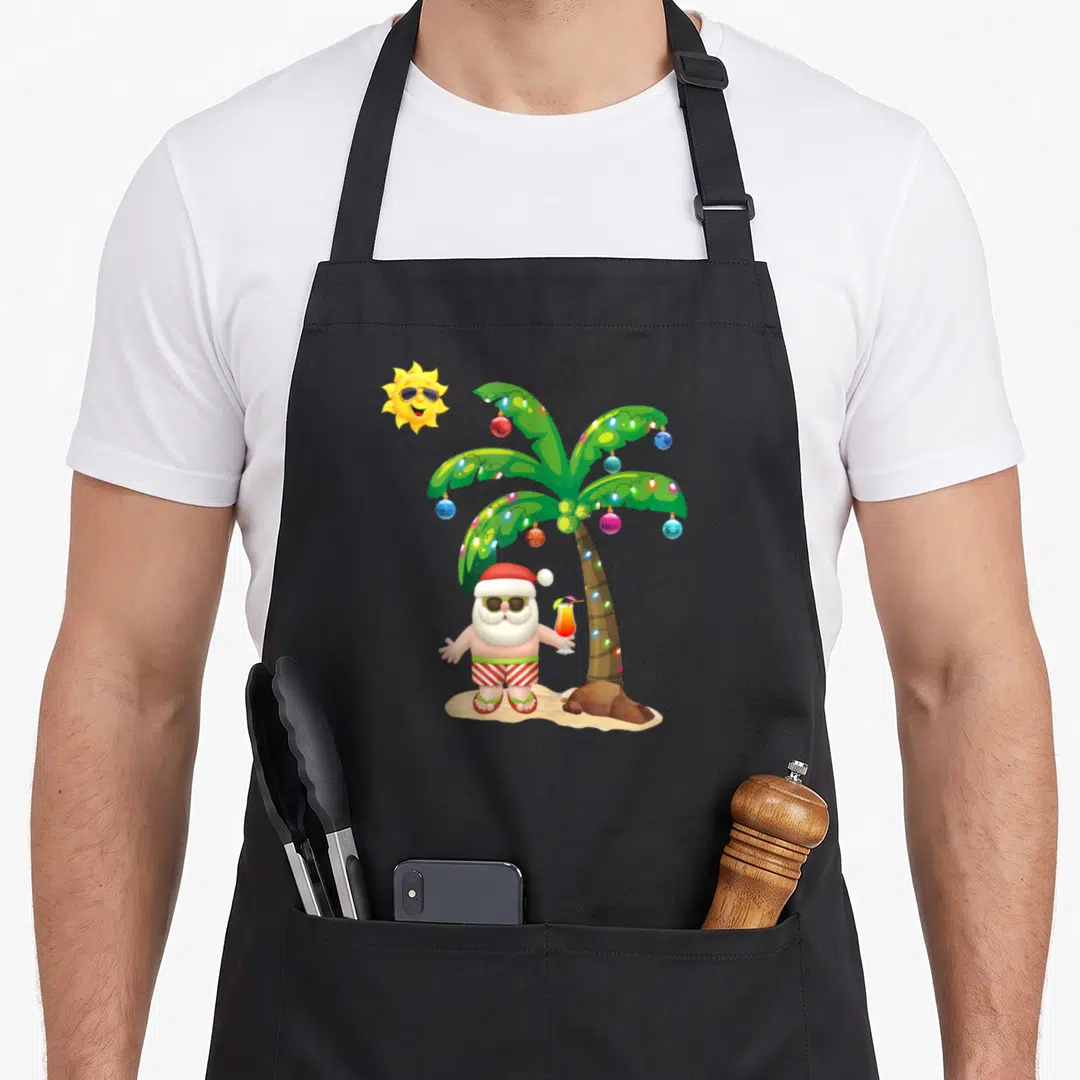 July santa claus apron - Cartoon Santa in red shorts holding a cocktail under a green palm tree with ornaments — shown on a black cotton canvas apron against a white background