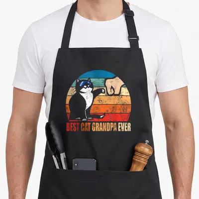 Granddad cat apron - Retro cat fist bumping human hand in striped circle with BEST CAT GRANDPA EVER text — shown on a black cotton canvas apron against a white background