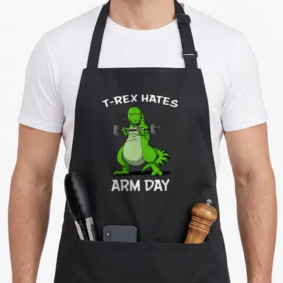 Bicep dinosaur apron - Green cartoon T-Rex lifting barbell with T-REX HATES ARM DAY text — shown on a black cotton canvas apron against a white background