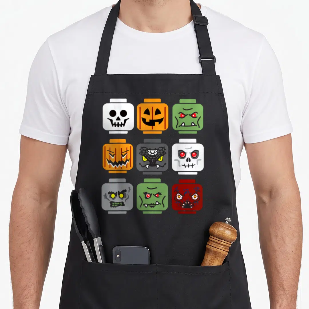 Halloween building apron - Nine toy blocks in a grid showing cartoon pumpkin, skull, and zombie faces in orange — shown on a black cotton canvas apron against a white background