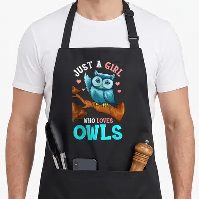 Cute owl apron - Blue cartoon owl perched on brown branch with pink hearts and white and pink text reading just a girl — shown on a black cotton canvas apron against a white background