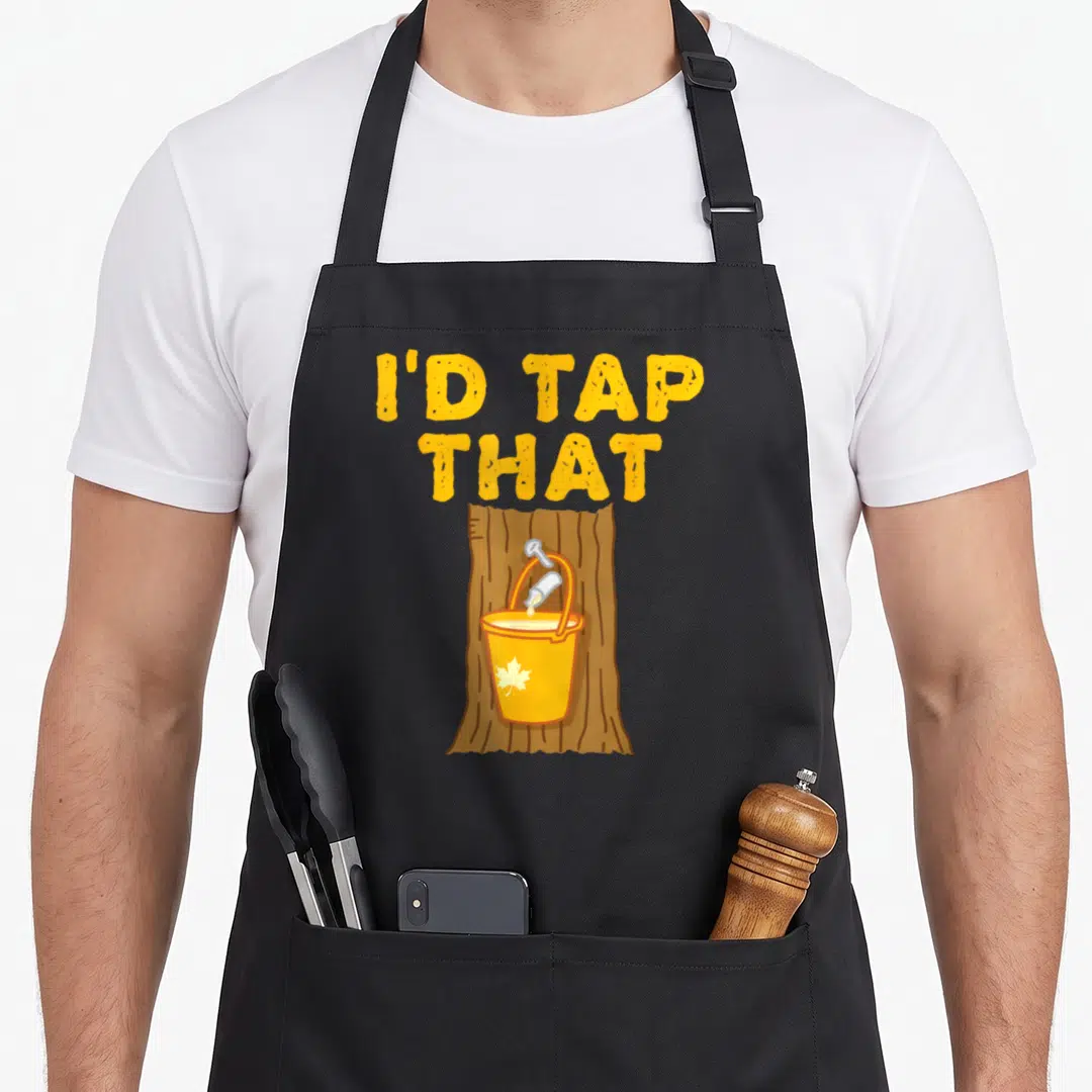 Tap tree apron - I'D TAP THAT text above yellow sap bucket with maple leaf on brown tree trunk — shown on a black cotton canvas apron against a white background