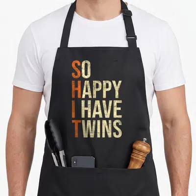 Twin mother's apron - Stacked text SO HAPPY I HAVE TWINS in orange and beige distressed typography — shown on a black cotton canvas apron against a white background