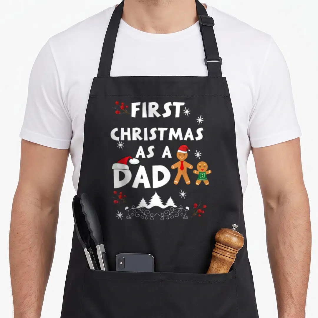 Funny xmas christmas aprons - FIRST CHRISTMAS AS A DAD text with gingerbread men, Santa hats, berries, snowflakes — shown on a black cotton canvas apron against a white background